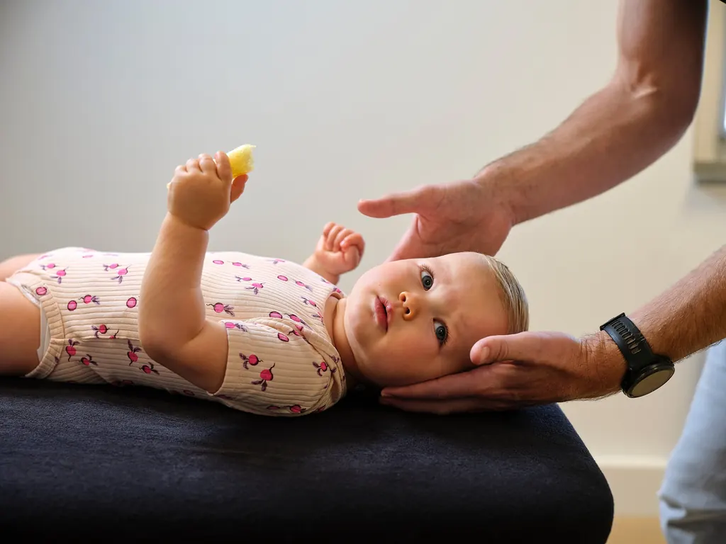 Een kindermanueel therapeut ondersteunt voorzichtig het hoofdje van een baby op de behandeltafel