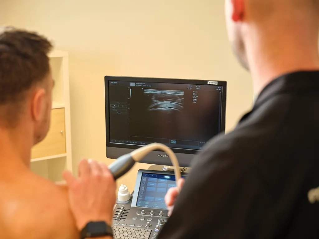 Een fysiotherapeut maakt een echo van de schouder van een cliënt; het beeld verschijnt op het scherm