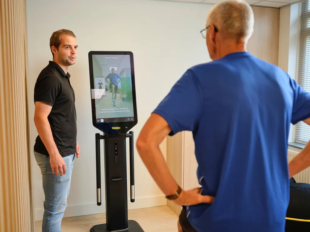 Een fysiotherapeut legt een balanstest uit terwijl een cliënt voor het meetscherm van de Technogym Checkup staat