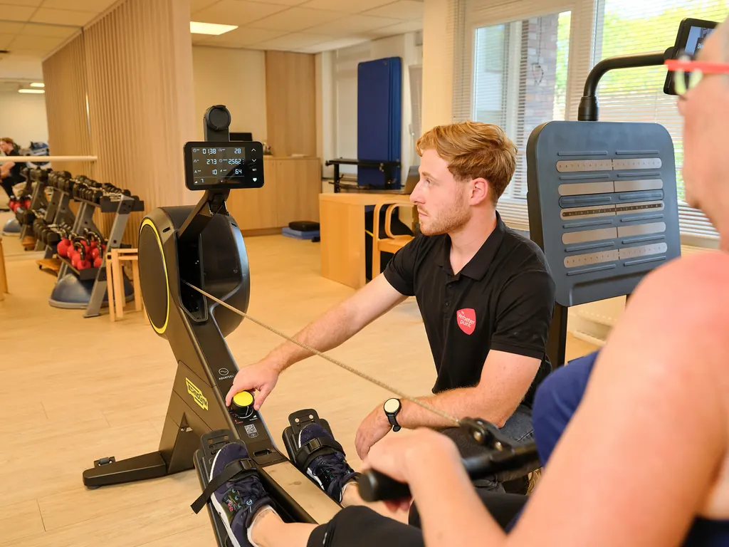 Een fysiotherapeut begeleidt een cliënt op een Technogym-roeitoestel met trainingsscherm